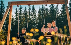 Riesenschaukel am Hirschi-Spielplatz, © Bergbahnen Semmering Hirschenkogel GmbH Drei Kinder schaukeln auf einer großen Schaukel im Freien, umgeben von Bäumen.