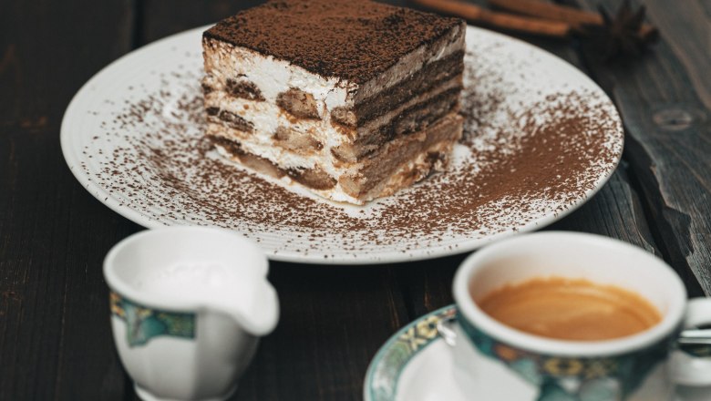 Hausgemachte Mehlspeisen, © cervodoro Ein Teller mit einem Stück Tiramisu, daneben eine Tasse Kaffee und ein Milchkännchen auf einem Holztisch.