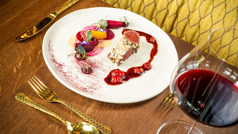 Ein Teller mit kunstvoll angerichtetem Rehrücken, Beilagen und einer Rotweinsoße, daneben ein Glas Rotwein auf einem Holztisch.