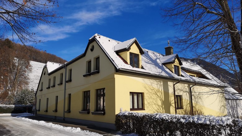 Hausansicht im Winter, © Gudrun Rath Gelbes Haus mit schneebedecktem Dach im Winter, blauer Himmel.