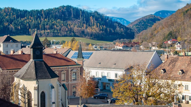 Stadtansicht von Gloggnitz mit Kapelle, Gebäuden und bewaldeten Hügeln im Hintergrund.