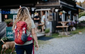 Frau mit rotem Rucksack und Hunde geht auf eine Berghütte zu.