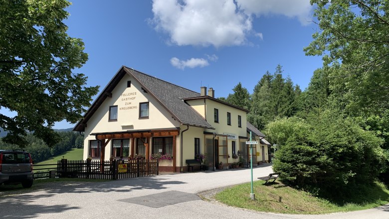 Ein gelbes Gasthaus mit der Aufschrift 'Polleres Gasthof zum Kreuzberg' umgeben von B&auml;umen und einem blauen Himmel.