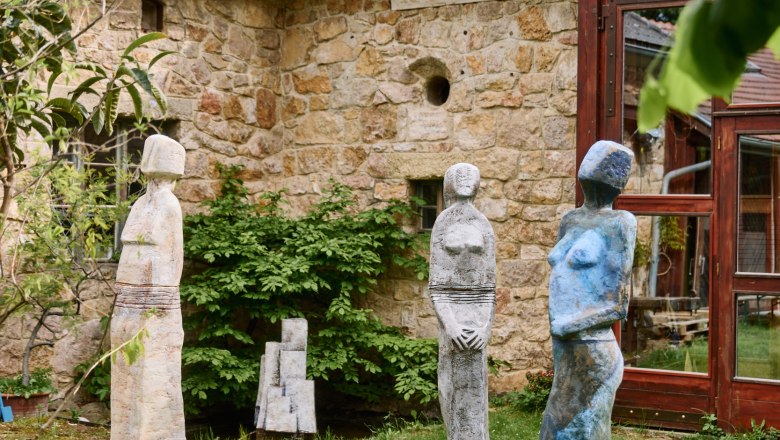 Drei bunte Skulpturen stehen auf einer Wiese vor einem alten Steingebäude