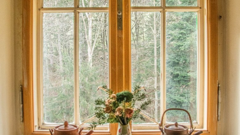 Fenster mit zwei Teekannen und einer Vase mit Blumen auf der Fensterbank, Wald im Hintergrund.