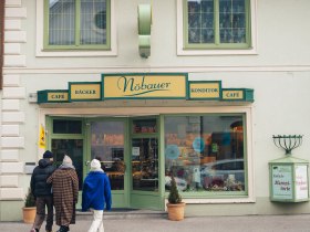 In der winterlichen Atmosphäre schlendern Besucher entspannt zur charmanten Konditorei, die mit ihren einladenden Fenstern und dem warmen Licht einladend wirkt. Der Duft frisch gebackener Leckereien zieht durch die kalte Luft und verspricht süße Genüsse, während die schneebedeckten Berge im Hintergrund eine malerische Kulisse bieten.