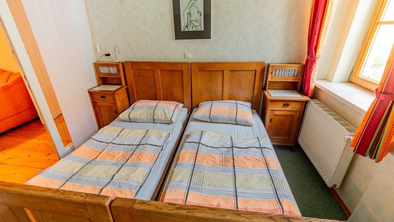 Ein Schlafzimmer mit zwei Einzelbetten, Holzm&ouml;beln und einem Fenster mit roten Vorh&auml;ngen.