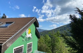 Grünes Haus mit Einhornbild an der Wand, umgeben von Bäumen und Bergen unter blauem Himmel.
