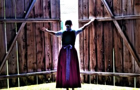 Eine Frau in traditioneller Kleidung &ouml;ffnet gro&szlig;e Holzt&uuml;ren nach au&szlig;en.