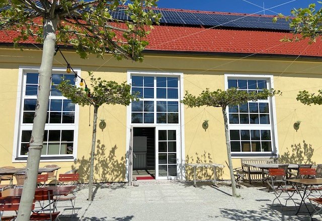 Gelbes Gebäude mit roten Dachziegeln, großen Fenstern und einem Biergarten mit Tischen und Stühlen im Freien.