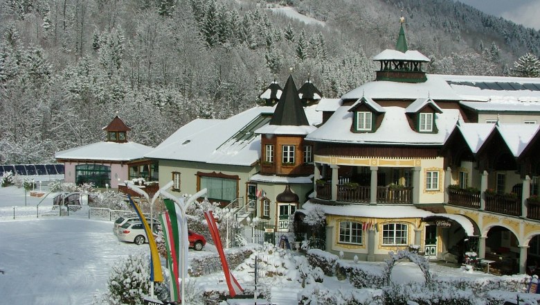 Raxalpenhof im Winter, © Scharfegger´s Raxalpen Resort Ein verschneites Hotel im alpinen Stil mit Türmen und Balkonen, umgeben von schneebedeckten Bäumen und Hügeln.
