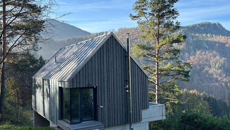 Modernes Holzhaus in bergiger Landschaft mit Bäumen und blauem Himmel.