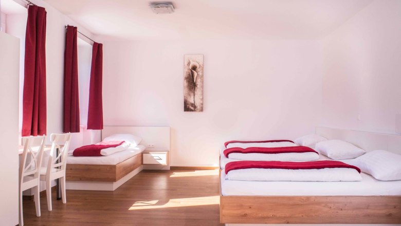 Helles Zimmer mit zwei Betten, roten Vorhängen und einem Bild an der Wand.