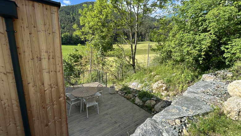 Terrasse mit Holztisch und Stühlen, umgeben von Bäumen und Wiese, mit Blick auf bewaldete Hügel.