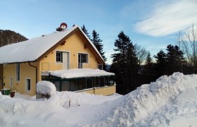 Ein gelbes Haus mit verschneitem Dach und Balkon, umgeben von B&auml;umen und Schnee.