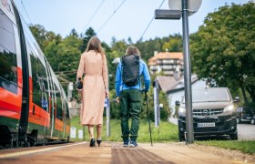 Zwei Personen am Bahnsteig, links der Zug und rechts ein Shuttlebus