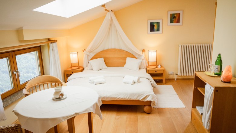 Gemütliches Doppelzimmer mit Holzmöbeln, großem Bett, Tisch und Stühlen, Fenster mit Aussicht und sanfter Beleuchtung.
