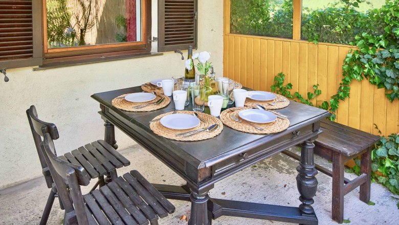 Gedeckter Tisch auf einer Terrasse mit vier Plätzen, Holzmöbeln und Pflanzen im Hintergrund.
