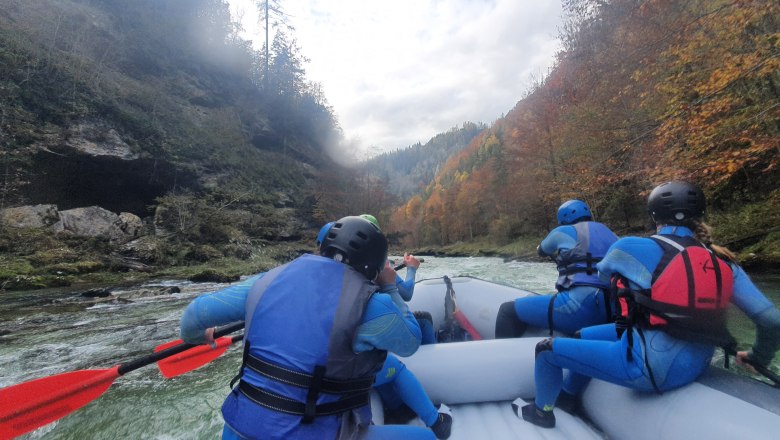 Gruppe beim Rafting auf einem Fluss, umgeben von herbstlichen B&auml;umen.