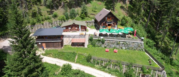Luftaufnahme der Potschacherh&uuml;tte in einem Waldgebiet mit umliegenden B&auml;umen und einer Terrasse mit Sonnenschirmen.