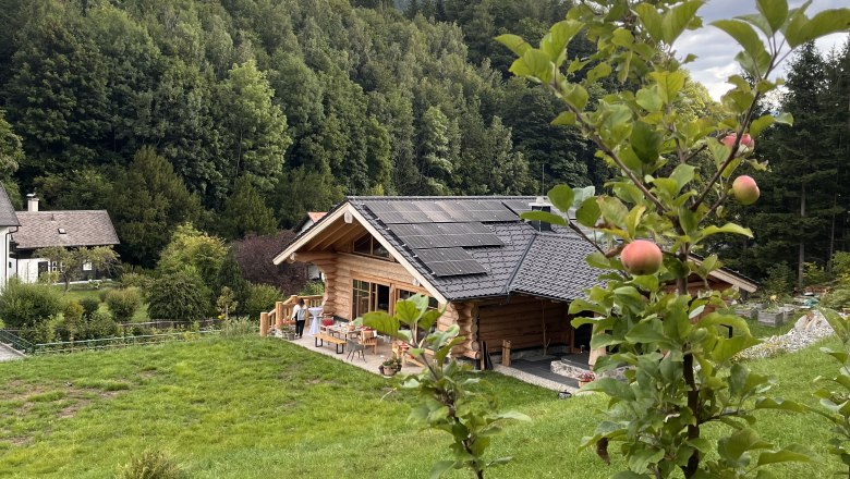 Ein Holzchalet mit Solarpanelen auf dem Dach, umgeben von gr&uuml;ner Natur und einem Apfelbaum im Vordergrund.