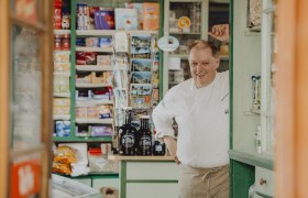 Ein Mann in Kochkleidung steht l&auml;chelnd in einem alten Laden mit Regalen voller Produkte.