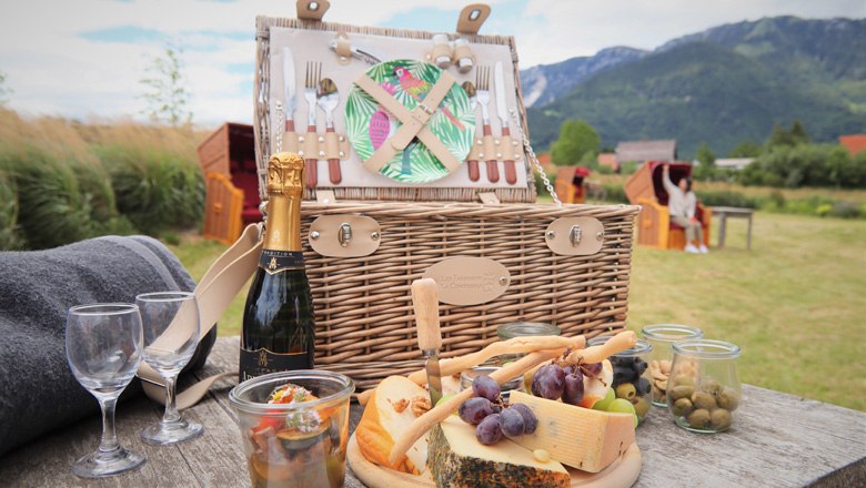 Picknickkorb mit K&auml;se, Trauben und Sektflasche auf einem Tisch in einer l&auml;ndlichen Umgebung mit Bergen im Hintergrund.