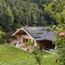 Ein Holzchalet mit Solarpanelen auf dem Dach, umgeben von gr&uuml;ner Natur und einem Apfelbaum im Vordergrund.