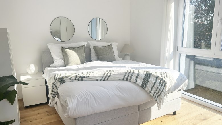 Helles Schlafzimmer mit Doppelbett, zwei runden Spiegeln an der Wand und gro&szlig;en Fenstern.