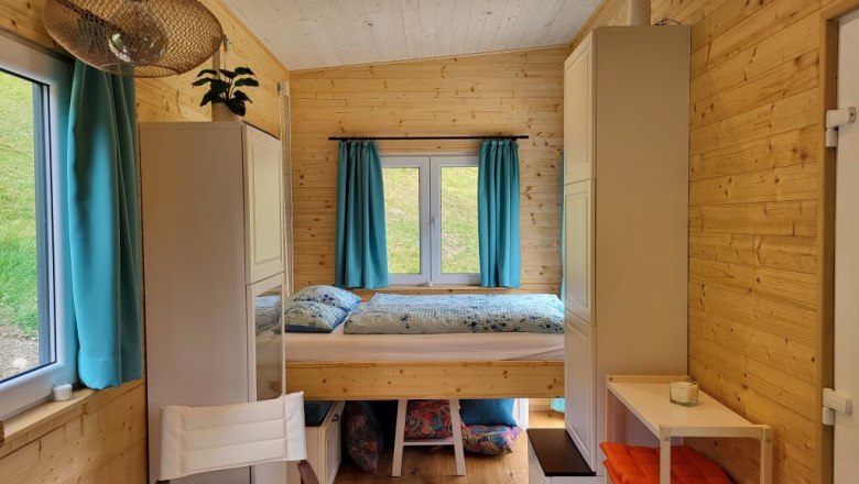Kleiner Raum mit Holzwänden, Hubbett, blauen Vorhängen und Regalen.