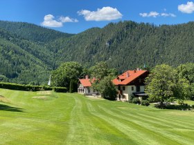 Ein Clubhaus mit rotem Dach in einer gr&uuml;nen, bergigen Landschaft unter blauem Himmel.