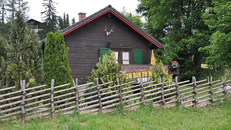 Eine rustikale Holzh&uuml;tte mit gr&uuml;nem Fensterladen und einem Holzzaun im Vordergrund, umgeben von B&auml;umen und Str&auml;uchern.