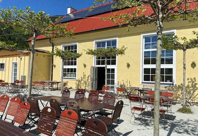 Außenbereich eines Restaurants mit gelbem Gebäude, roten Stühlen und Tischen unter Bäumen.