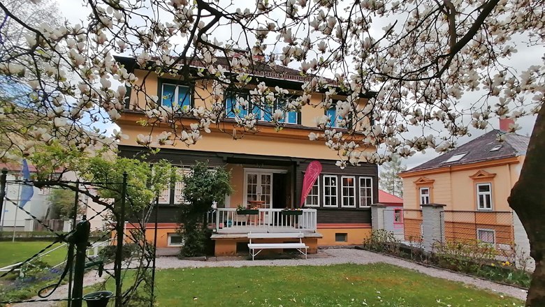 Ein zweistöckiges Haus mit gelber Fassade und weißen Fenstern, umgeben von blühenden Bäumen.