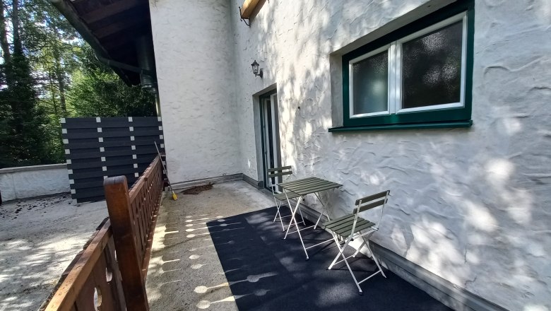 Terrasse einer Ferienwohnung mit Tisch und St&uuml;hlen, umgeben von B&auml;umen.