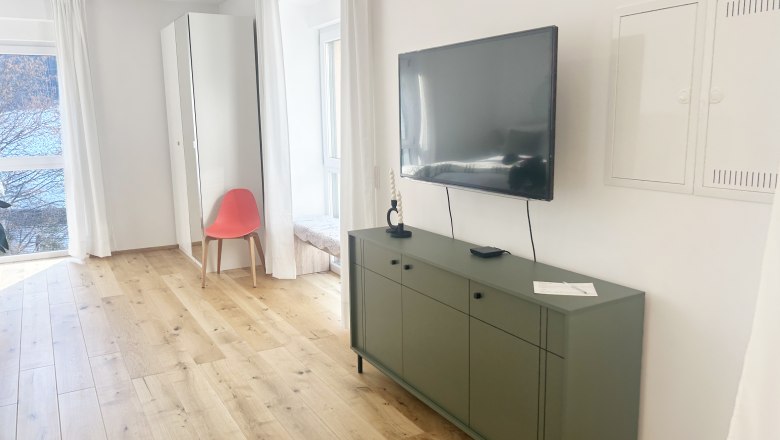 Modernes Wohnzimmer mit Holzboden, gr&uuml;nem Sideboard, Wandfernseher und rotem Stuhl vor einem Spiegel.
