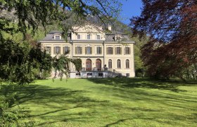 Ein großes, elegantes Herrenhaus mit symmetrischer Fassade und mehreren Fenstern, umgeben von grüner Wiese und Bäumen.