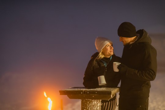 Zwei Personen stehen im Wintergewand an einem Stehtisch mit einer Tasse in der Hand und im Hintergrund ein Lagerfeuer