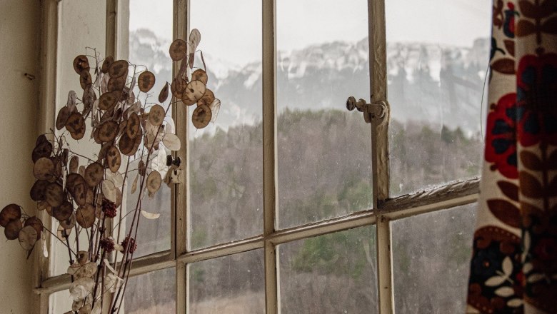 Blick aus einem Fenster mit Berglandschaft im Hintergrund, Vase mit Trockenblumen im Vordergrund.