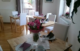 Ein heller Frühstücksraum mit Holztischen, weißen Stühlen und Blumenarrangements auf den Tischen.