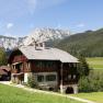 Ein traditionelles Holzhaus in einer bergigen Landschaft mit Wiesen und W&auml;ldern im Hintergrund.
