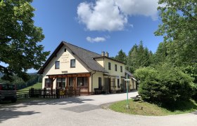 Ein gelbes Gasthaus mit der Aufschrift 'Polleres Gasthof zum Kreuzberg' umgeben von Bäumen und einem blauen Himmel.