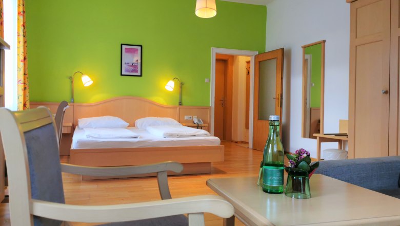 Ein gemütliches Doppelzimmer mit grün gestrichener Wand, Doppelbett, Tisch, Stühlen und Wasserflasche.