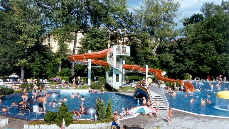 Ein Freibad mit vielen Menschen, einer Wasserrutsche und B&auml;umen im Hintergrund.