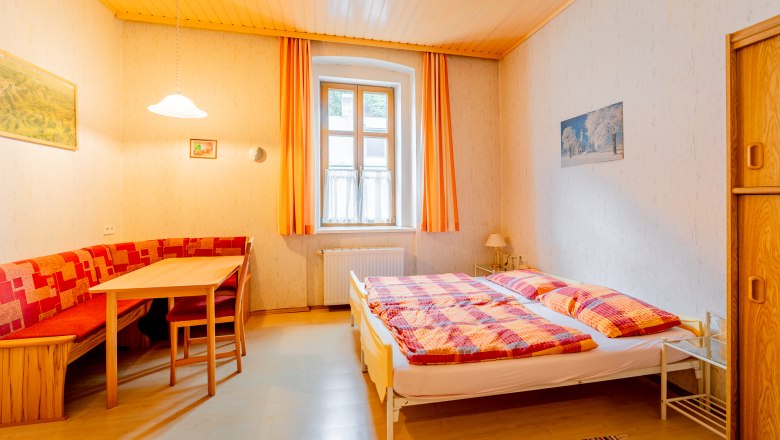 Gem&uuml;tliches Schlafzimmer mit Doppelbett, Holzm&ouml;beln und Esstisch. Helle Vorh&auml;nge und warme Farben dominieren den Raum.