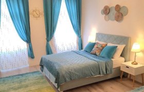 Ein gem&uuml;tliches Schlafzimmer mit blauen Vorh&auml;ngen, einem Doppelbett und dekorativen Kissen. Zwei Nachttischlampen beleuchten den Raum.