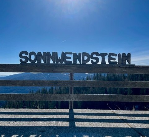 Gro&szlig;buchstaben mit 'Sonnwendstein' vor sonnigem Himmel und Berglandschaft.