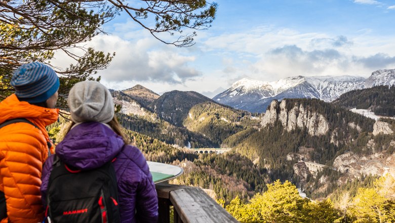 Zwei Personen betrachten eine Berglandschaft mit Viadukt.