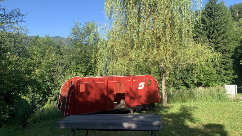 Rote Spielzeuglokomotive im Freien neben einem Baum und einer Holzbank.