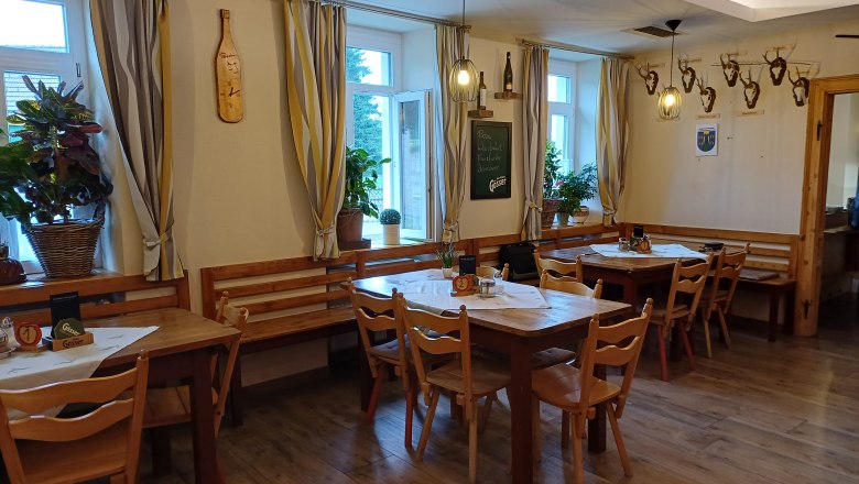 Innenansicht eines gemütlichen Gastraums mit Holztischen und Stühlen, dekoriert mit Pflanzen und Hirschgeweihen an der Wand.